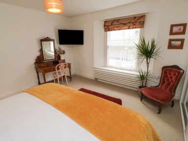 A bedroom with a bed covered with an orange blanket a wooden dressing table with a mirror and chair a wall-mounted TV a window with blinds a red armchair a potted plant and two framed pictures at Lands House in Appledore
