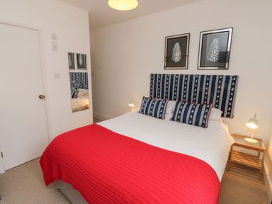 A bedroom with a bed with red blanket and blue patterned cushions a bedside table with a lamp and two framed pictures on the wall at Lands House in Appledore