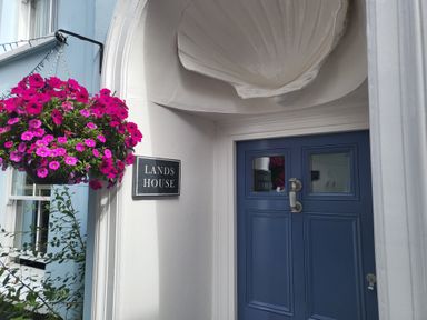 A blue door with silver handle under a white shell-shaped overhang next to a hanging basket of pink flowers at Lands House in Appledore
