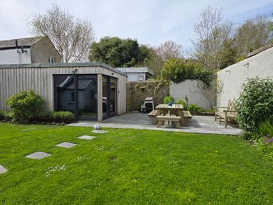 A backyard with a wooden picnic table a bench a barbecue and a small building with glass doors at Lands House in Appledore