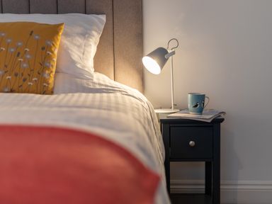 A bed with white sheets and a yellow pillow next to a black nightstand with a lamp a mug and magazines at Lands House in Appledore