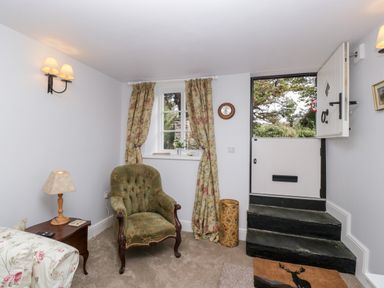 A small room with a green armchair, floral curtains, a side table with a lamp, and a half door with steps at The Old Thatch in Porlock