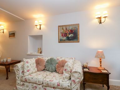 A sitting area with a floral sofa, round cushions, a wooden side table with a lamp and small decor, wall sconces and floral painting at The Old Thatch in Porlock