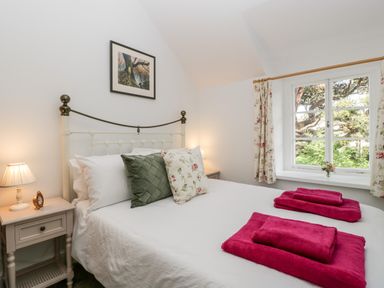 A bedroom with a double bed white bedding green and floral pillows pink towels on the bed bedside tables with lamps a framed picture on the wall and a window with floral curtains at The Old Thatch in Porlock