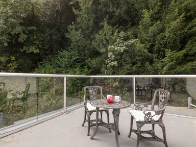 An outdoor patio with a metal table and two chairs and a red teapot and white mugs on the table surrounded by trees at The Old Thatch in Porlock