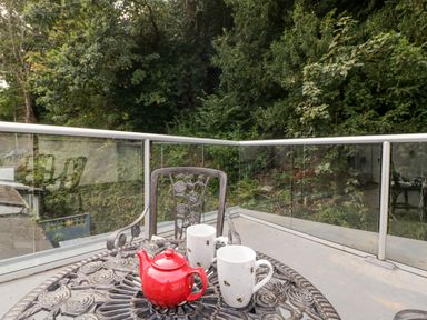 An outdoor patio with a black metal table and chairs a red teapot and two white mugs on the table surrounded by glass railing with trees in the background at The Old Thatch in Porlock