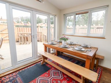 A dining area with a wooden table set with plates and glasses with benches on a red patterned rug next to French doors and a window with blinds at Gloccamaura in Bovey Tracey