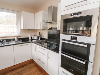 A kitchen with white cabinets black countertops a double oven electric stovetop and a window with blinds at Gloccamaura in Bovey Tracey