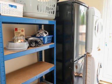 A utility room with a blue storage shelving unit containing pet supplies and laundry baskets next to a black fridge and a stacked white washer and dryer at Gloccamaura in Bovey Tracey
