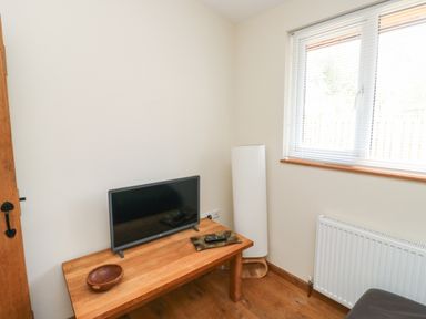 A small room with a wooden table holding a flat screen TV and a wooden bowl next to a tall white lamp near a window with blinds and a radiator below at Gloccamaura Bovey Tracey