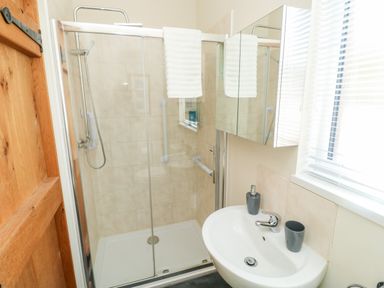 A bathroom with a wooden door shower enclosure with glass doors and a tiled wall a white sink with a faucet a soap dispenser and a cup on the sink and a mirrored cabinet above the sink a window with blinds