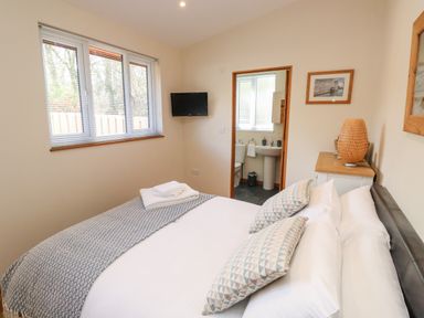 A bedroom with a bed pillows a patterned blanket a window a wall-mounted TV and an open door to an en suite bathroom at Gloccamaura in Bovey Tracey