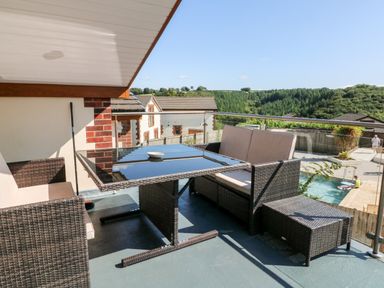 A balcony with wicker furniture and a glass table overlooking houses and a pool at Hazelwood Lodge in High Bickington