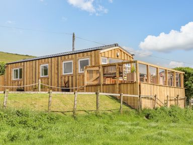 A wooden cabin with a fenced deck and outdoor furniture on a grassy hill at Caban Cwtch The Cosy Cabin in Llanaelhaearn