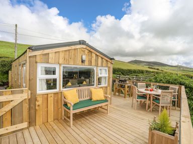 A wooden deck with outdoor seating and a dining table next to a wooden cabin surrounded by green hills at Caban Cwtch The Cosy Cabin in Llanaelhaearn