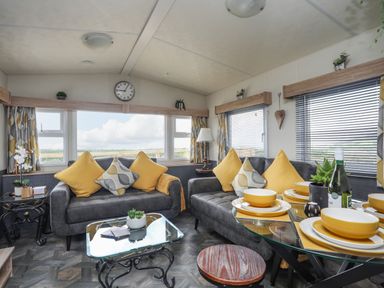 A living room with two grey sofas with yellow and patterned cushions a glass coffee table and a dining table set with yellow dishes at Caban Cwtch (The Cosy Cabin) in Llanaelhaearn