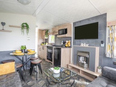 A small kitchen and dining area with wooden cabinets a stove a microwave a dining table with yellow dishes and stools a fireplace with a television above it and a glass coffee table at Caban Cwtch The Cosy Cabin in Llanaelhaearn