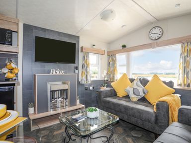 A living room with a gray sofa with yellow and patterned cushions a glass coffee table TV above a fireplace and windows with patterned curtains at Caban Cwtch The Cosy Cabin in Llanaelhaearn