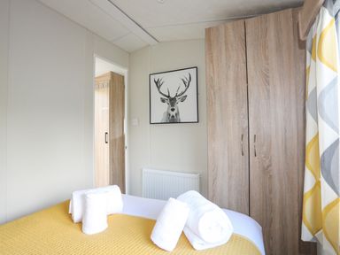 A bedroom with a bed covered in a yellow blanket and white towels on it a wardrobe a framed picture of a deer and patterned curtains at Caban Cwtch (The Cosy Cabin) in Llanaelhaearn