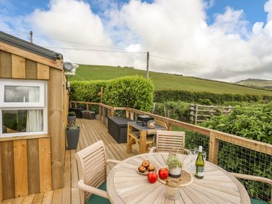 An outdoor wooden deck with a round table, chairs, and a scenic countryside view at Caban Cwtch in Llanaelhaearn