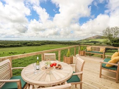 A wooden deck with table and chairs overlooking green fields and hills at Caban Cwtch The Cosy Cabin in Llanaelhaearn
