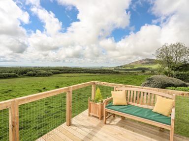 A wooden deck with a bench and two yellow cushions overlooking green fields and a hill at Caban Cwtch The Cosy Cabin in Llanaelhaearn