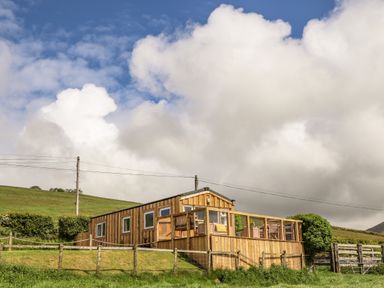 A small wooden cabin with a fenced deck on a grassy hill at Caban Cwtch The Cosy Cabin in Llanaelhaearn