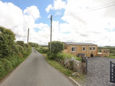 A narrow country road next to a wooden cabin with a gravel driveway and greenery at Caban Cwtch The Cosy Cabin in Llanaelhaearn
