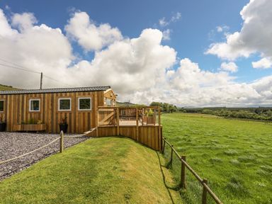 A wooden cabin with a deck and gravel path next to a grassy field and wooden fence at Caban Cwtch The Cosy Cabin in Llanaelhaearn