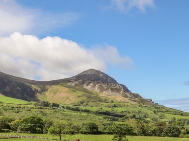 A mountain with green fields and trees under a blue sky with clouds at Caban Cwtch The Cosy Cabin in Llanaelhaearn