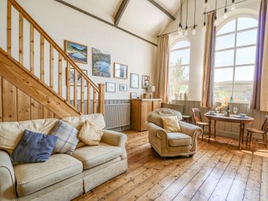 A sitting area with a beige sofa and armchair near a wooden staircase and a dining table with chairs by large windows at St Marks School House Nant-Glas near Rhayader