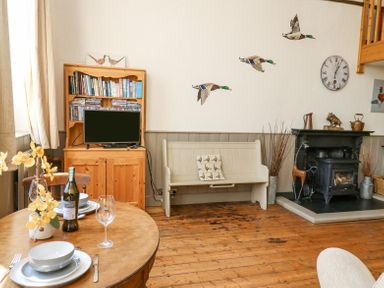 A dining table with dishes and wine bottle next to a wooden cabinet with books and television a white bench a fireplace and wall decorations at St Marks School House Nant-Glas near Rhayader