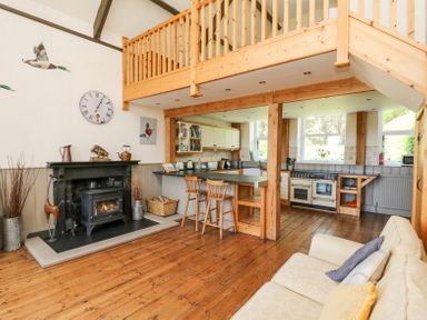 An open kitchen and living area with wooden floor a white sofa a wood-burning stove and a loft balcony at St Marks School House in Nant-Glas near Rhayader