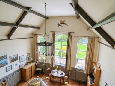 A dining area with a round wooden table set for two under a hanging chandelier in a room with large arched windows and wooden beams at St Marks School House Nant-Glas near Rhayader