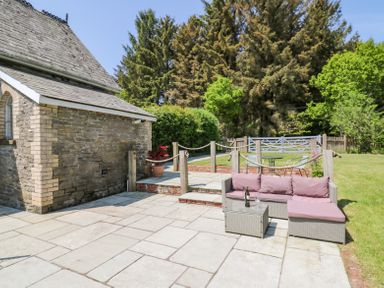 An outdoor patio area with a stone building wooden posts rope fencing a wicker sofa with pink cushions and a grass lawn with trees at St Marks School House Nant-Glas near Rhayader