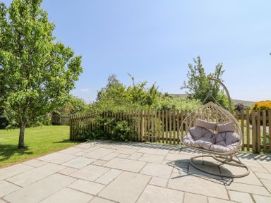 An outdoor patio with a hanging chair and cushions next to a wooden picket fence and green trees at St Marks School House in Nant-Glas near Rhayader