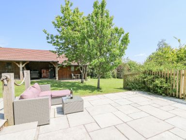 An outdoor patio with a wicker sofa and table on stone tiles next to a lawn with trees and a wooden shed at St Marks School House in Nant Glas near Rhayader
