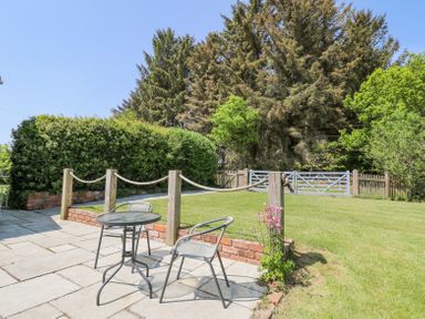 An outdoor patio with a small round table and two chairs overlooking a grassy yard with a rope fence and dense trees at St Marks School House Nant-Glas near Rhayader