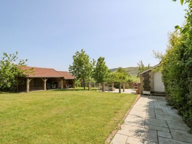 A grassy backyard with trees a stone patio area wooden outbuildings and a paved walkway at St Marks School House in Nant-Glas near Rhayader