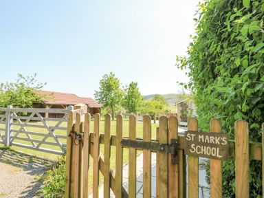A wooden gate with a sign reading St Marks School leading to a yard with grass, trees, and buildings at St Marks School House in Nant-Glas near Rhayader