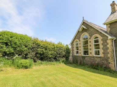 A lawn with bushes next to a stone building with arched windows at St Marks School House in Nant-Glas near Rhayader