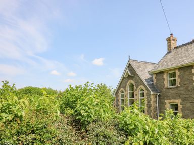 A stone house with arched windows partially obscured by green bushes at St Marks School House in Nant-Glas near Rhayader