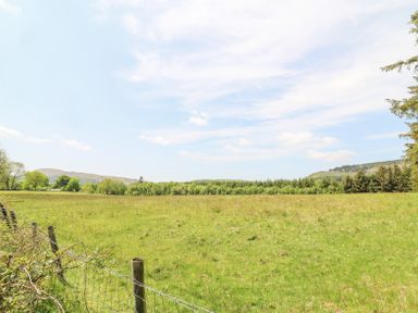 A grassy field with a wire fence and trees in the background under a partly cloudy sky at St Marks School House in Nant-Glas near Rhayader