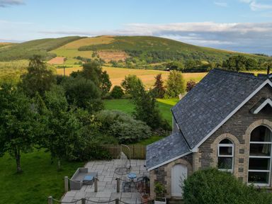 A stone house with a slate roof and patio furniture in a garden with trees and hills in the background at St Marks School House in Nant-Glas near Rhayader