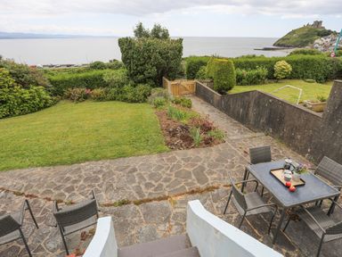 A garden with a stone patio and outdoor table with chairs overlooking the sea and a hill with a castle at Llys Owen in Criccieth