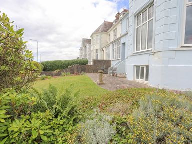 A garden with grass and plants next to a light blue building with large windows and patio furniture at Llys Owen in Criccieth