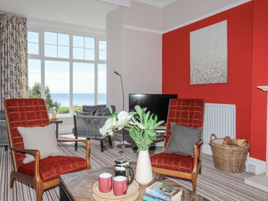 A living room with two red chairs a wooden coffee table with flowers cups and books and a window with a sea view at Llys Owen in Criccieth
