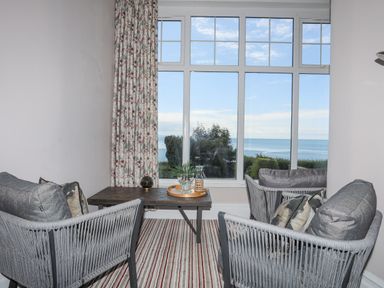 A seating area with two gray armchairs a wooden table and a large window showing a sea view at Llys Owen in Criccieth
