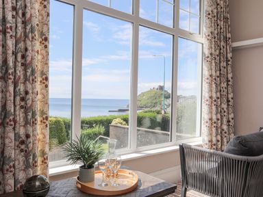 A room with large windows showing a sea view a tray with glasses and plant on a table and a chair by the window at Llys Owen in Criccieth