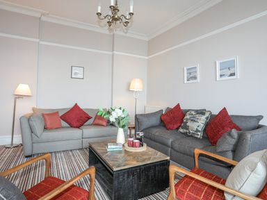 A living room with two grey sofas with red cushions a wooden coffee table with flowers and books and two wooden armchairs with red cushions at Llys Owen in Criccieth